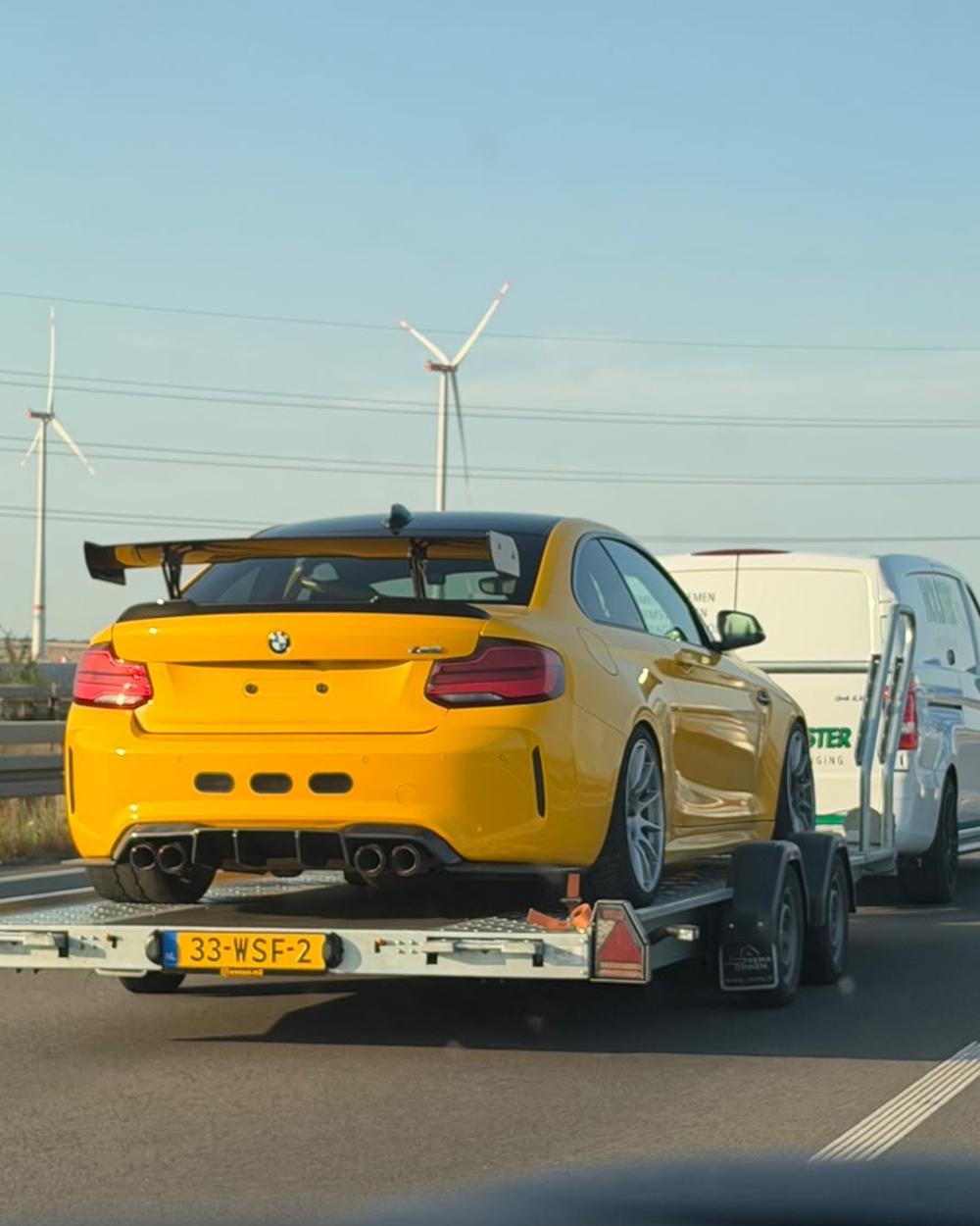 Gele BMW M2 met achtervleugel op de aanhanger, onderweg over de snelweg